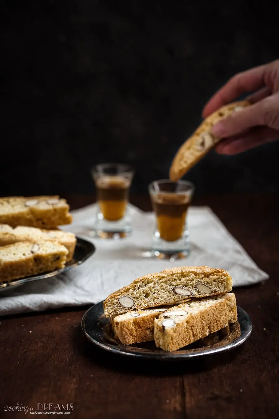 3 biscotti on a tray and in the background a hand dipping the cookie it in vin santo