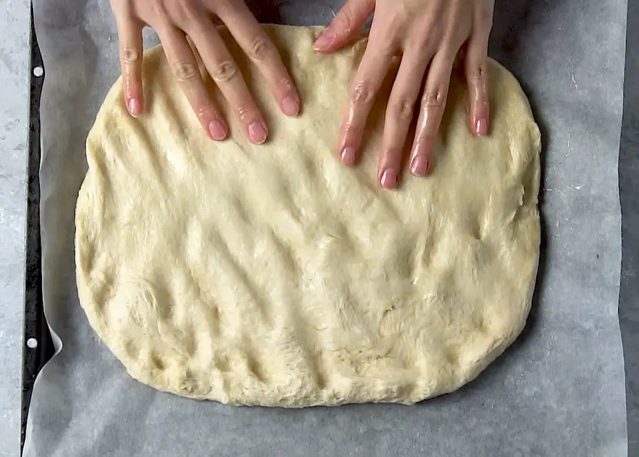 hands stretching the focaccia dough on a lined baking sheet