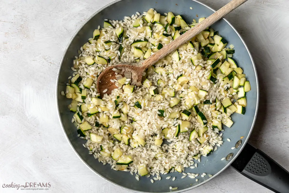 rice is added to the pan with zucchini and mixed with a wooden spoon