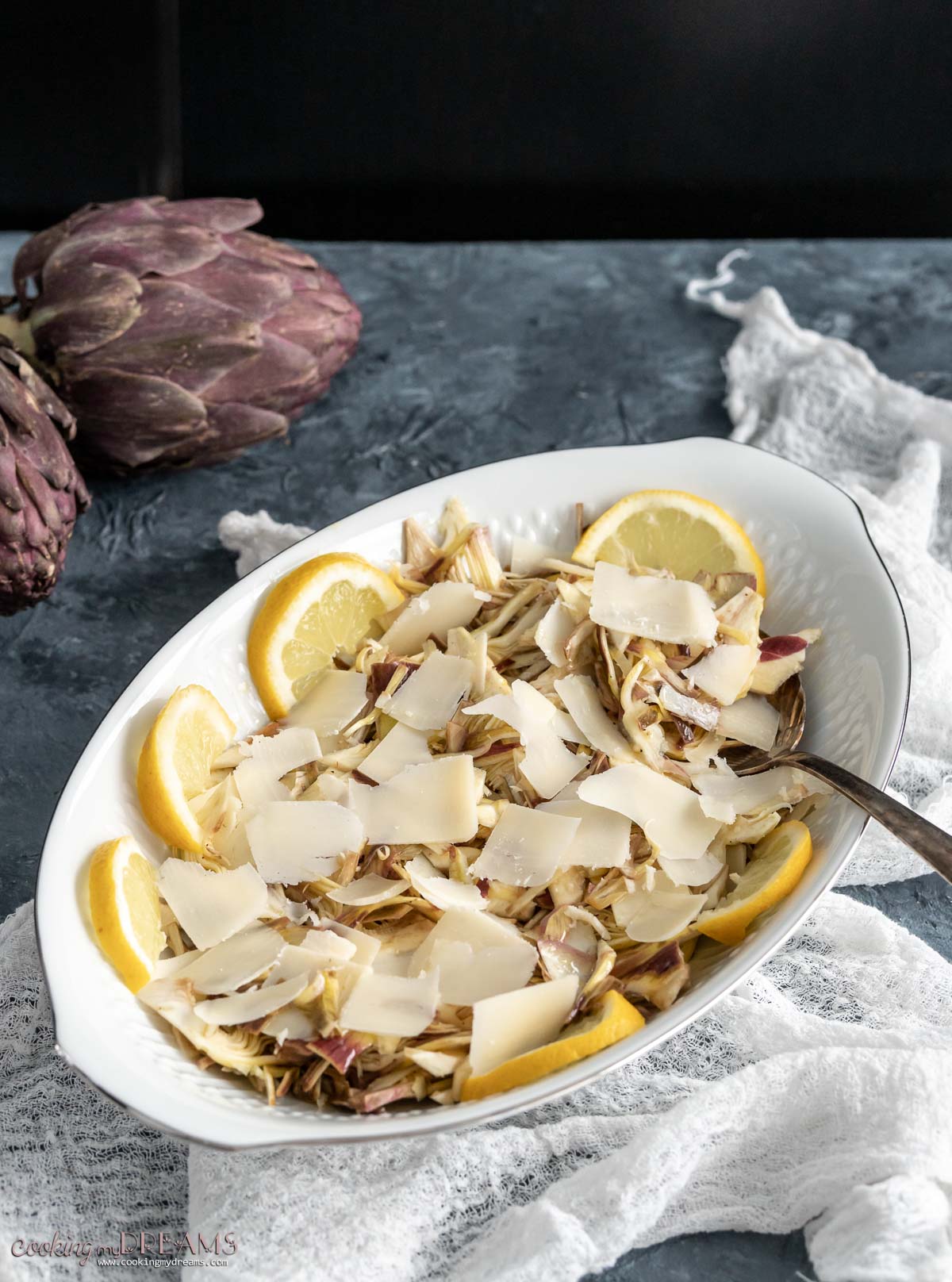shaved artichoke salad with parmigiano cheese and lemon slices on an oval platter with a serving spoon