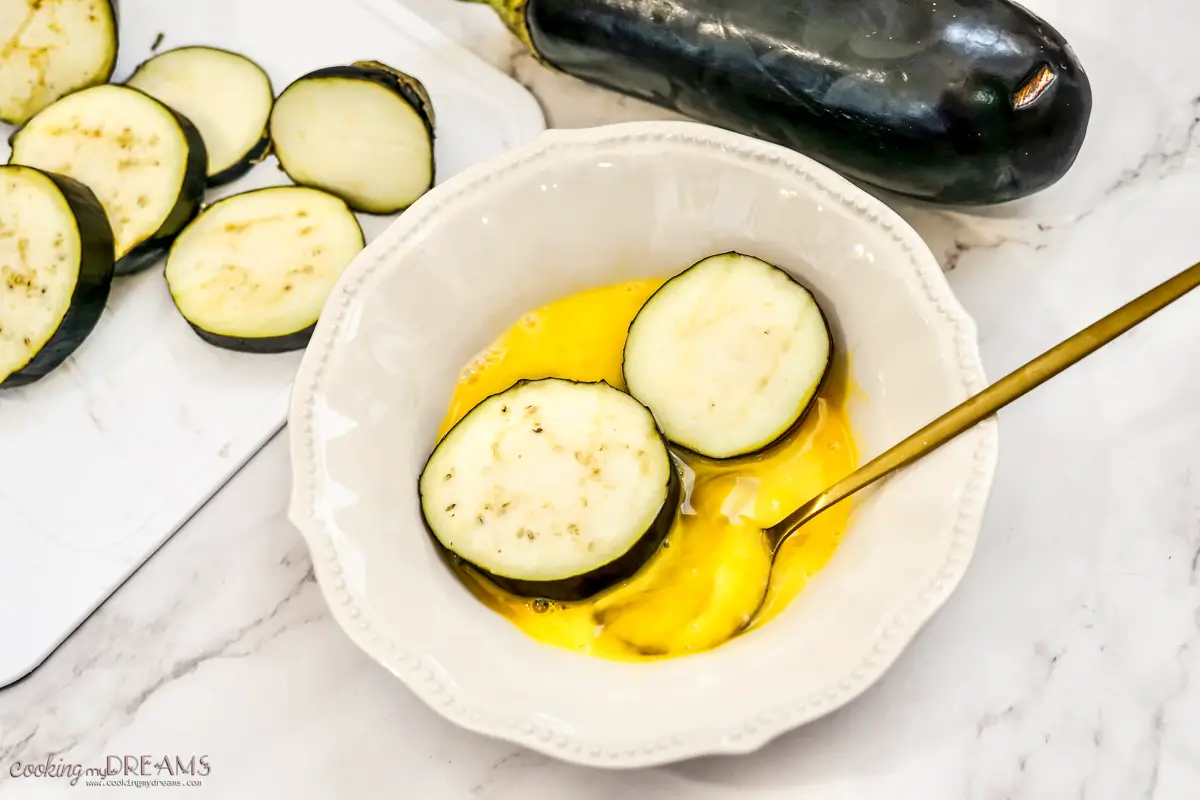 eggplant slices being coated in egg wash in a plate with a spoon