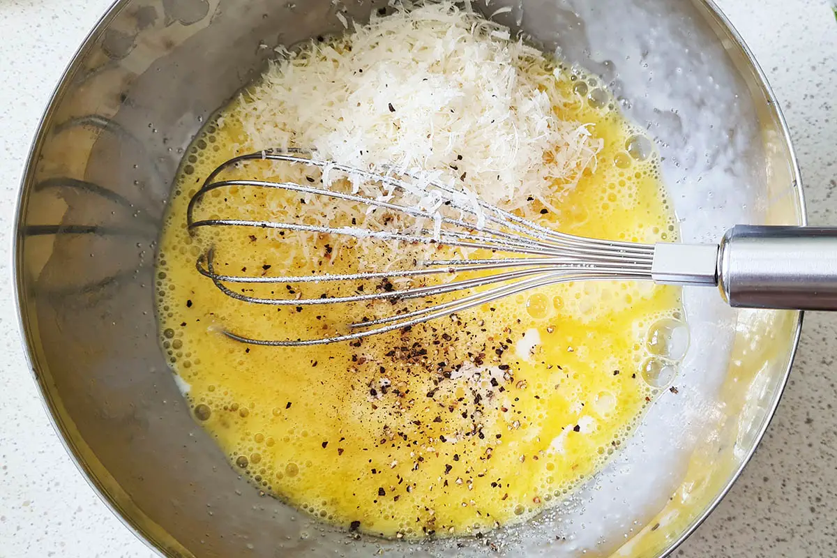 a whisk in a bowl with eggs, parmesan, and seasonings.