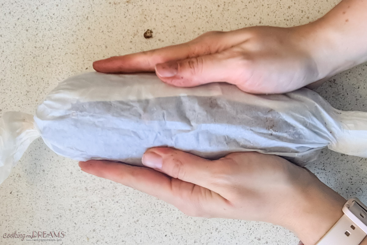 hands holding a chocolate salami wrapped in parchment paper.