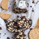 chocolate salami sliced on paper next to biscuits and hazelnuts.