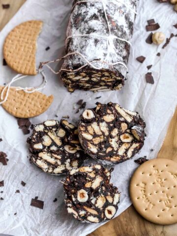chocolate salami sliced on paper next to biscuits and hazelnuts.