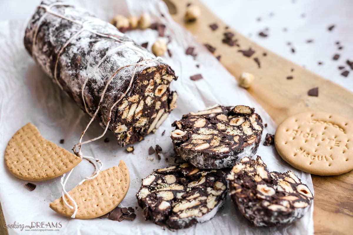 chocolate salami sliced on paper next to biscuits and hazelnuts.