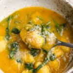 Spoon lifting pumpkin gnocchi soup with spinach.