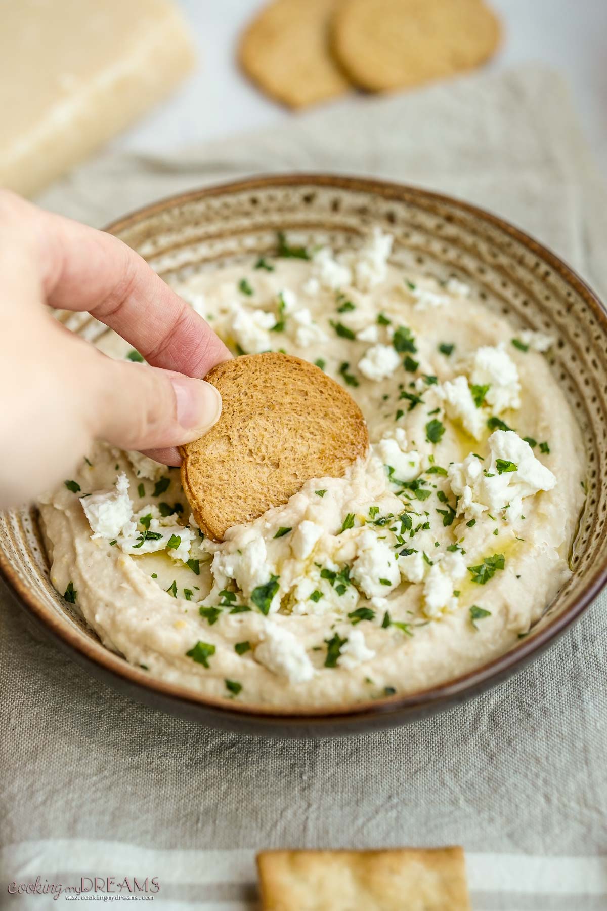 hand holding a cracker taking a bit of white bean dip from the bowl.