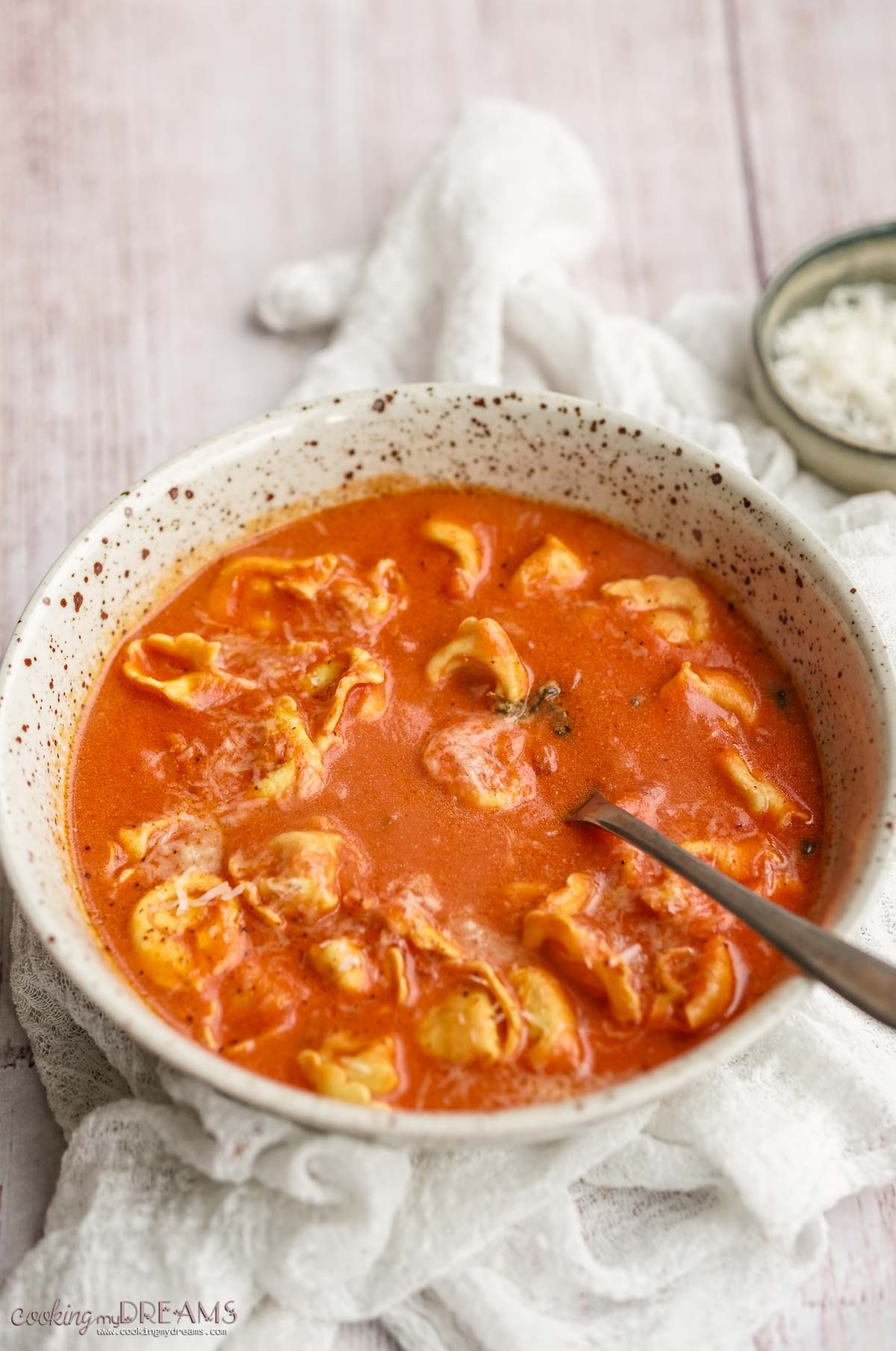 spoon inside a bowl of tomato tortellini soup on a white table.