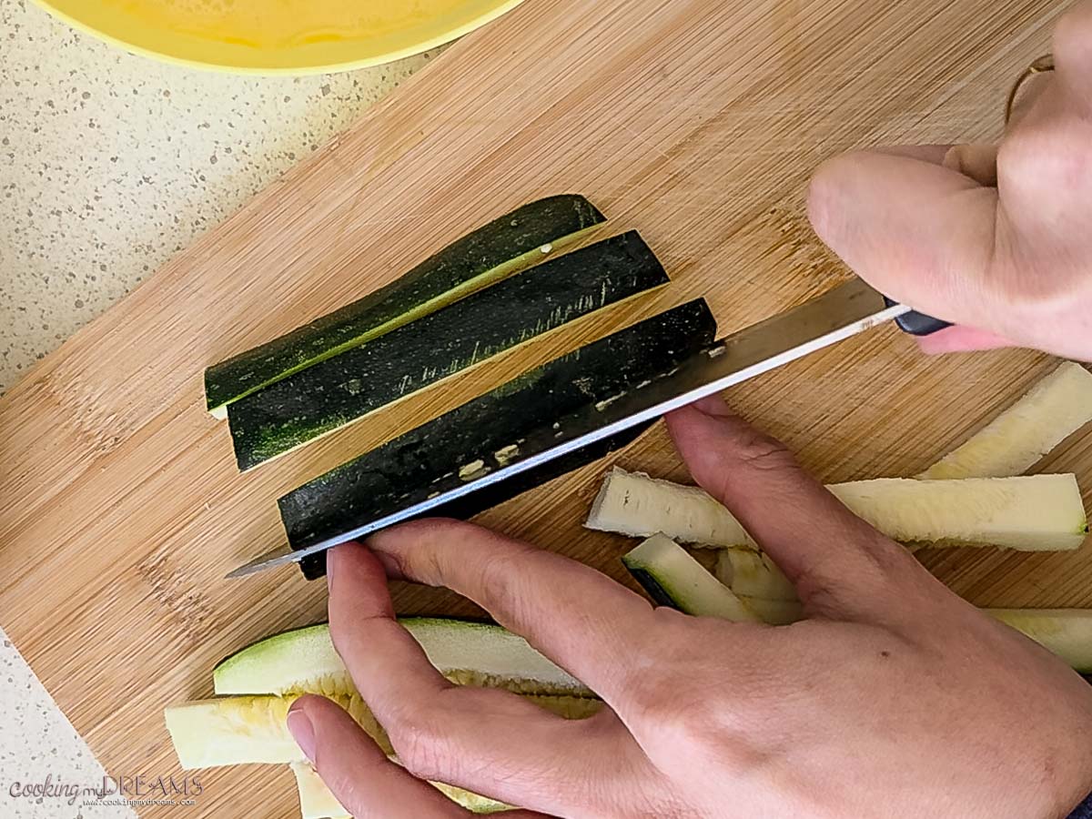 hands cutting zucchini into sticks on a wooden board.