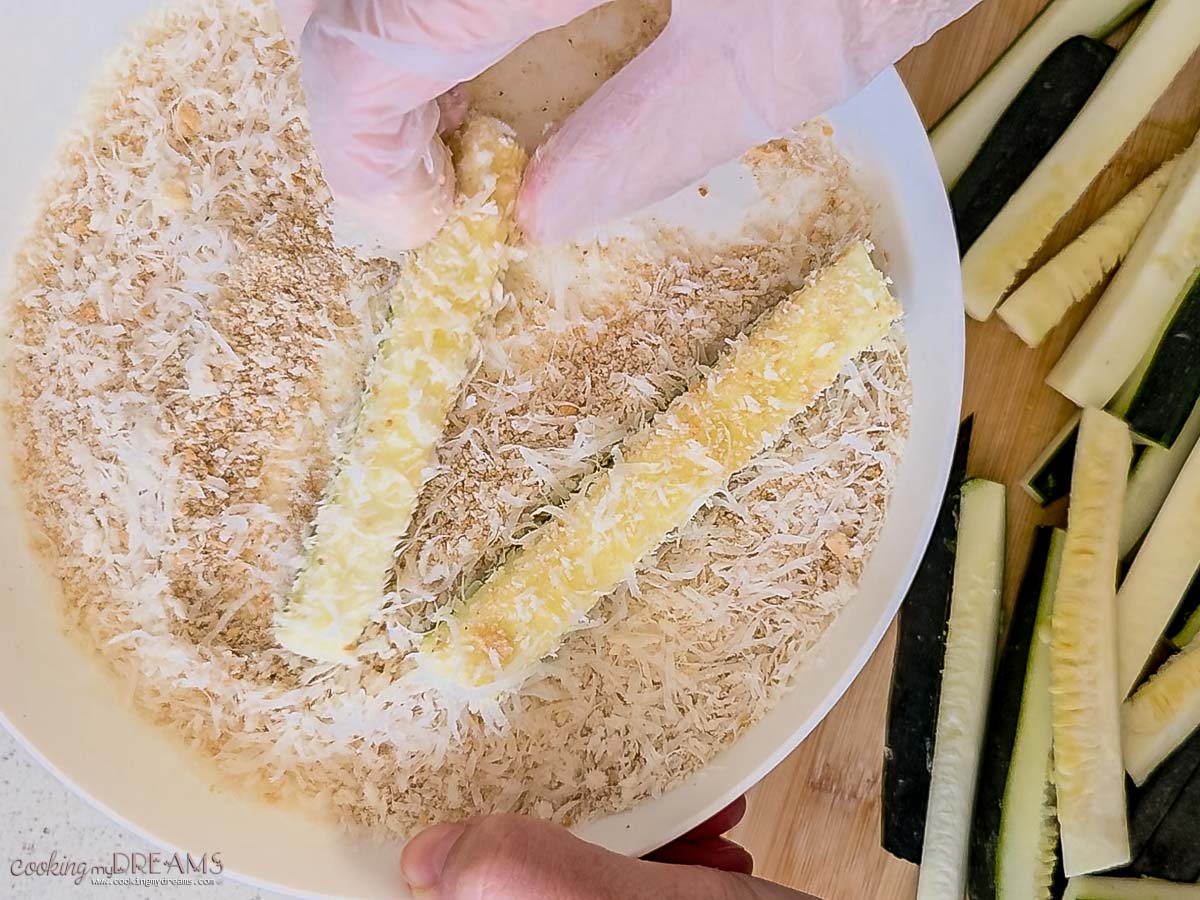 hand dipping the zucchini sticks into breadcrumbs mix.