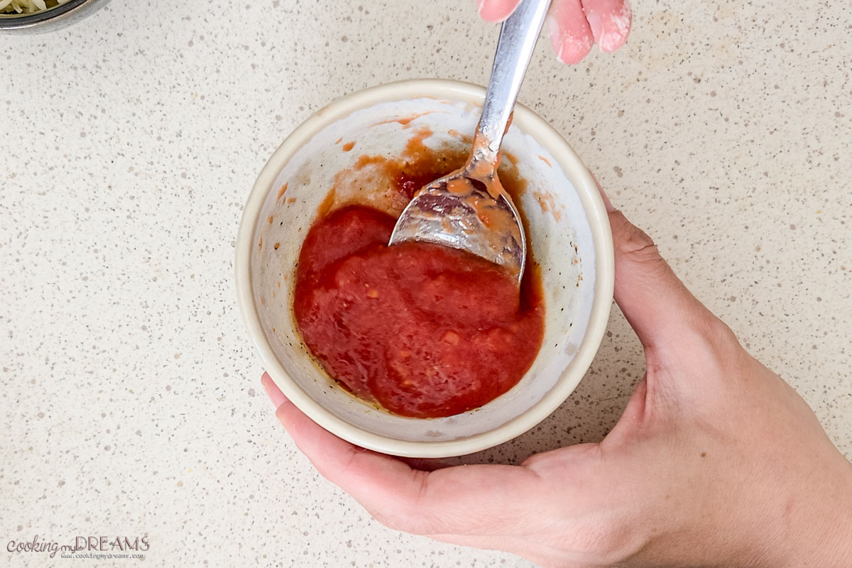 hands mixing pizza sauce in a small bowl with a spoon.